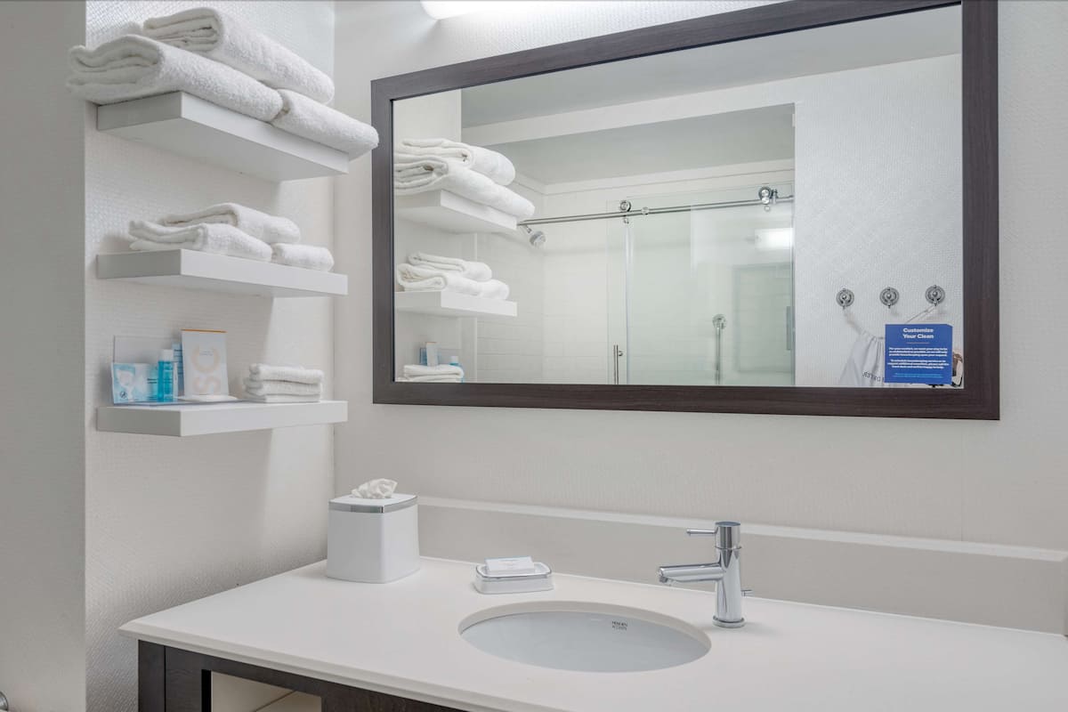 A bathroom with a large mirror, white shelves with folded towels, a sink, and a shower area.
