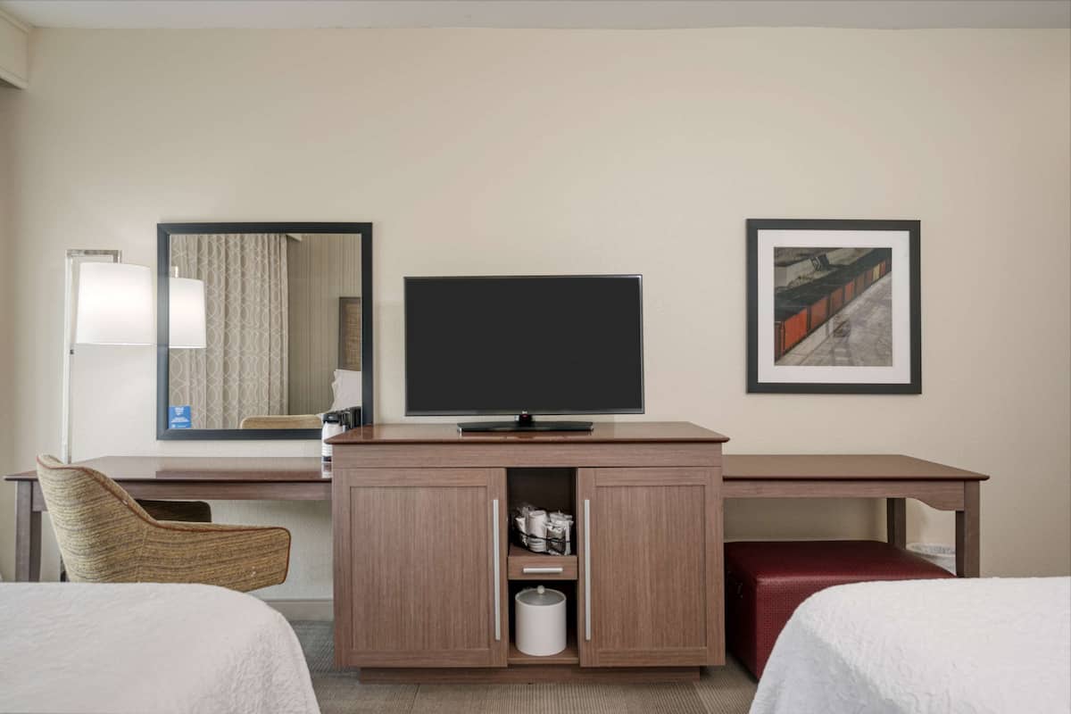 A hotel room with a bed, a TV on a wooden cabinet, a chair, and a desk with a lamp.