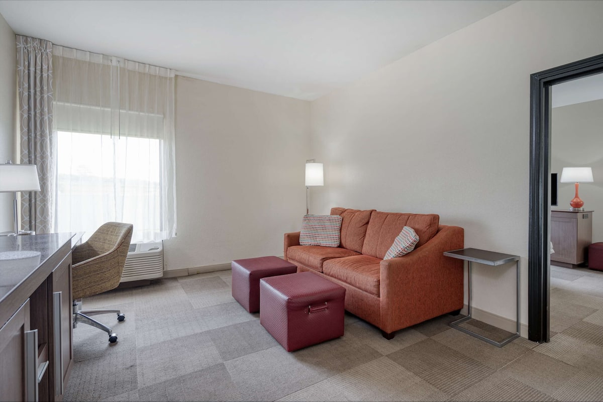 A hotel room with a sofa, two ottomans, a desk with a chair, a lamp, and a large mirror.
