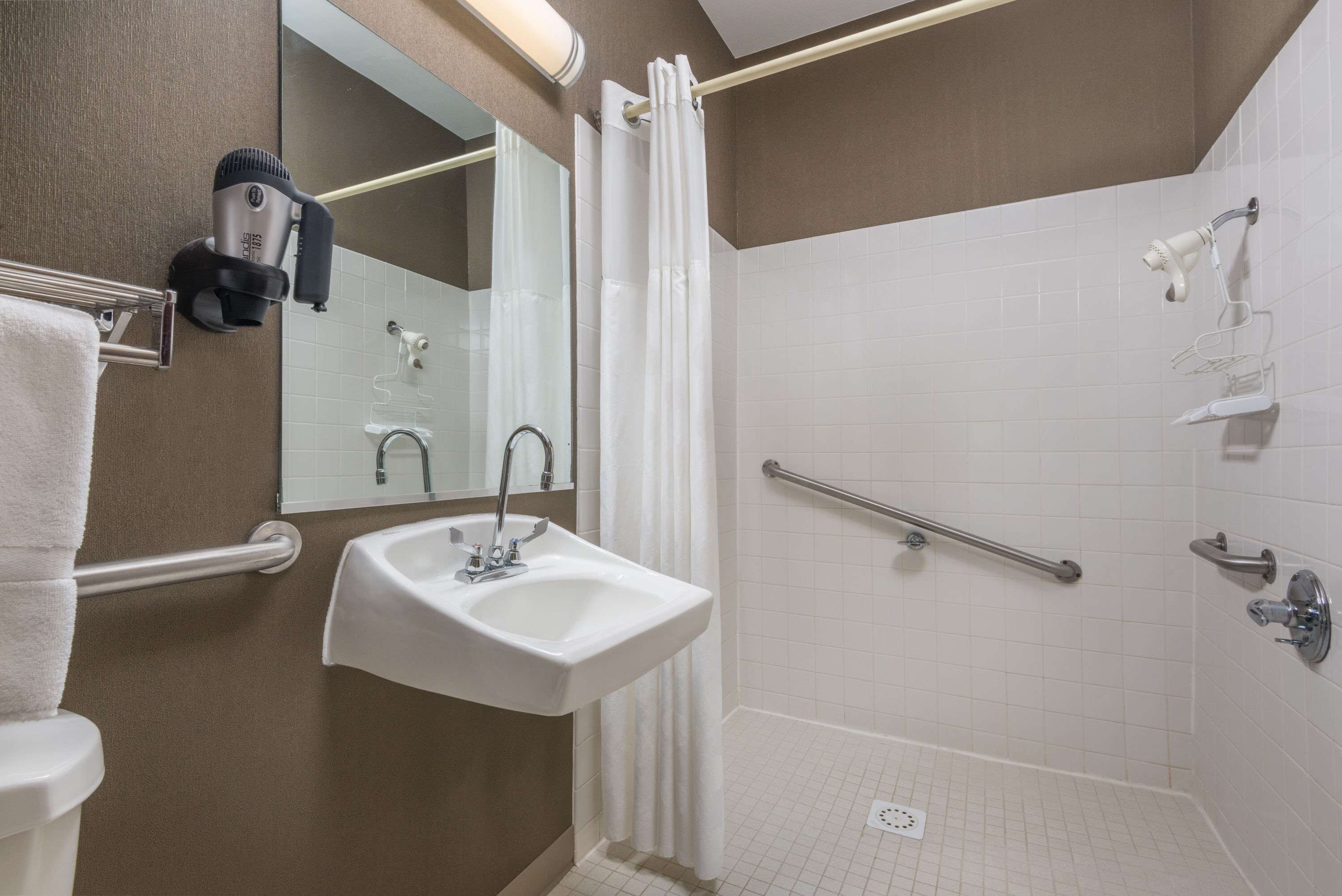A bathroom with a sink, mirror, towel rack, shower with grab bars, and a hairdryer mounted on the wall.