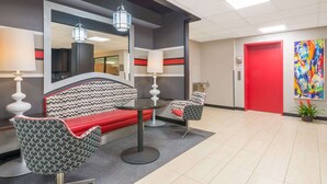 A modern lobby with a red door, a large mirror, a sofa, two patterned chairs, and a small plant.