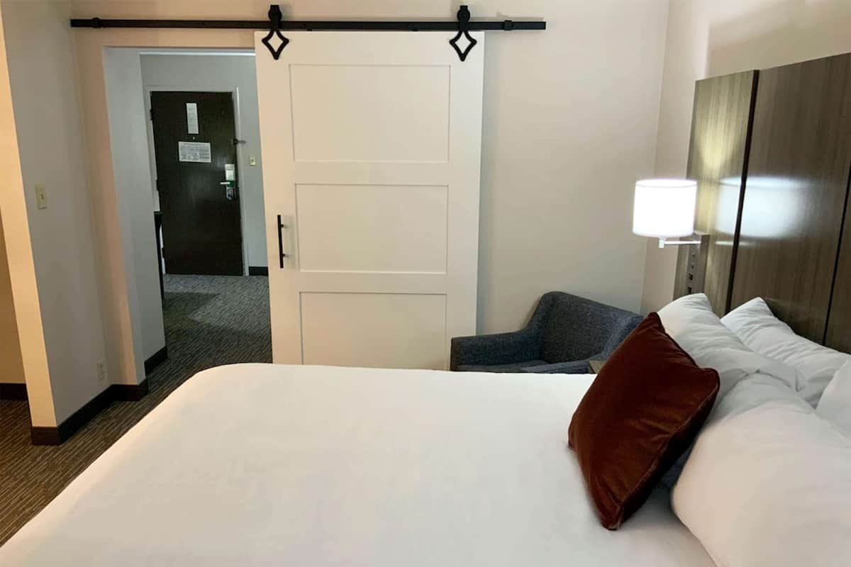 A hotel room with a large bed, a desk, and a chair. There is a door leading to another room.