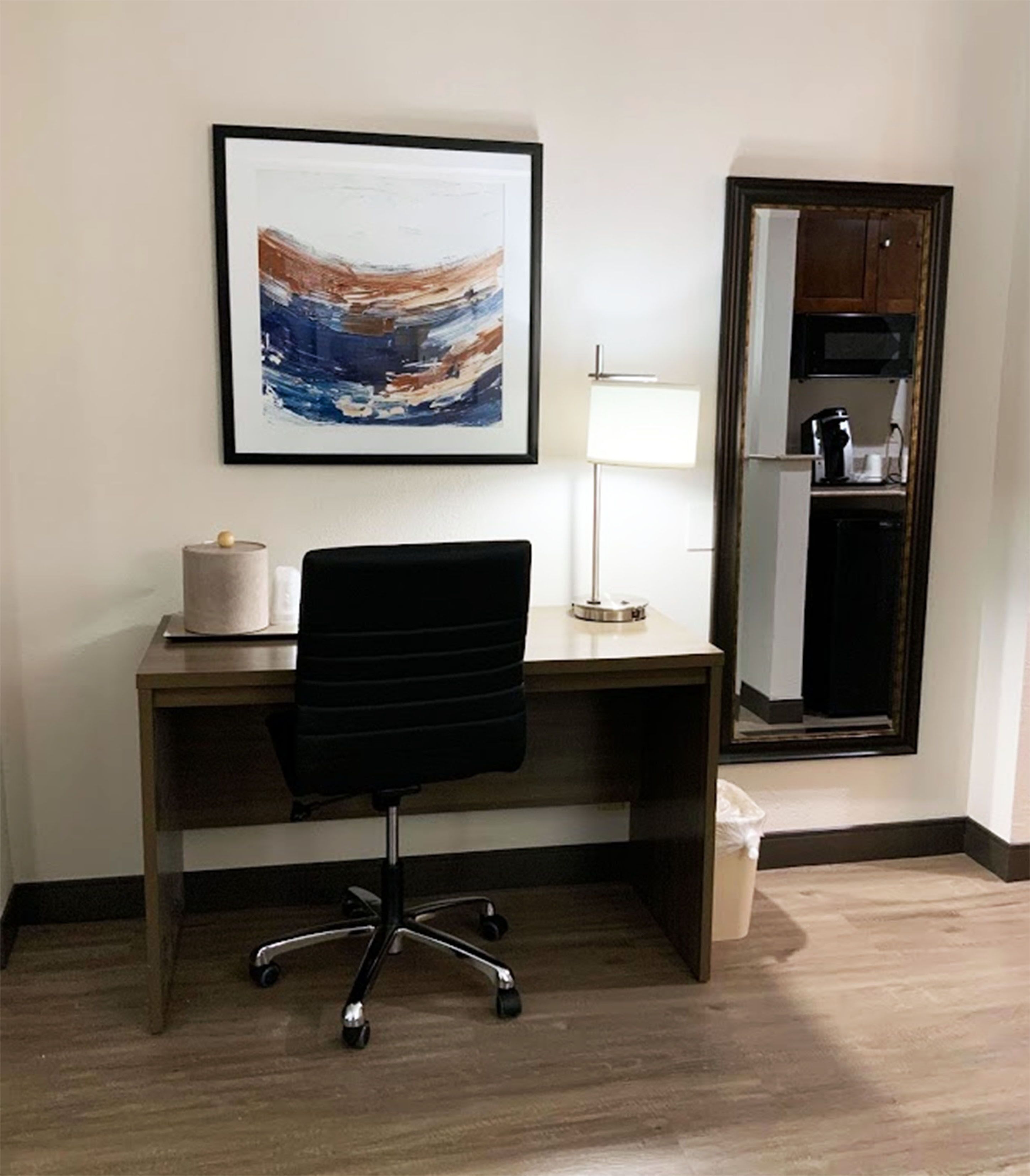 A hotel room with a wooden desk, a black office chair, a framed abstract painting, a floor lamp, and a mirror reflecting a kitchenette.