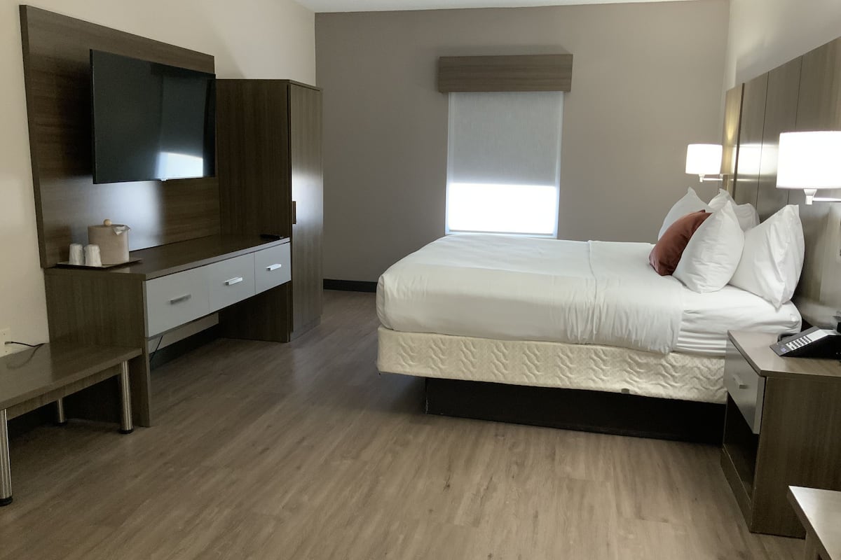 A hotel room with a bed, a TV mounted on the wall, a bedside table, a desk, and a window with blinds.