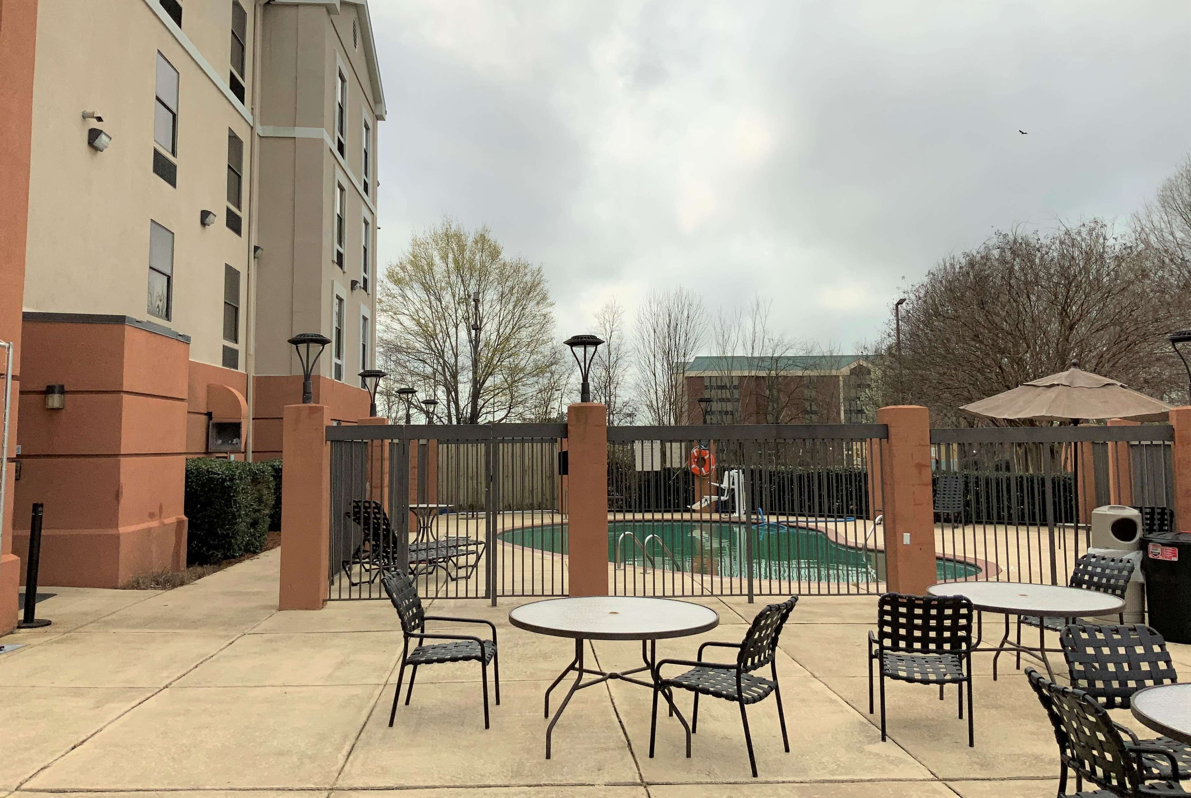 Un patio al aire libre con mesas y sillas, una piscina y un área cercada con un edificio al fondo.