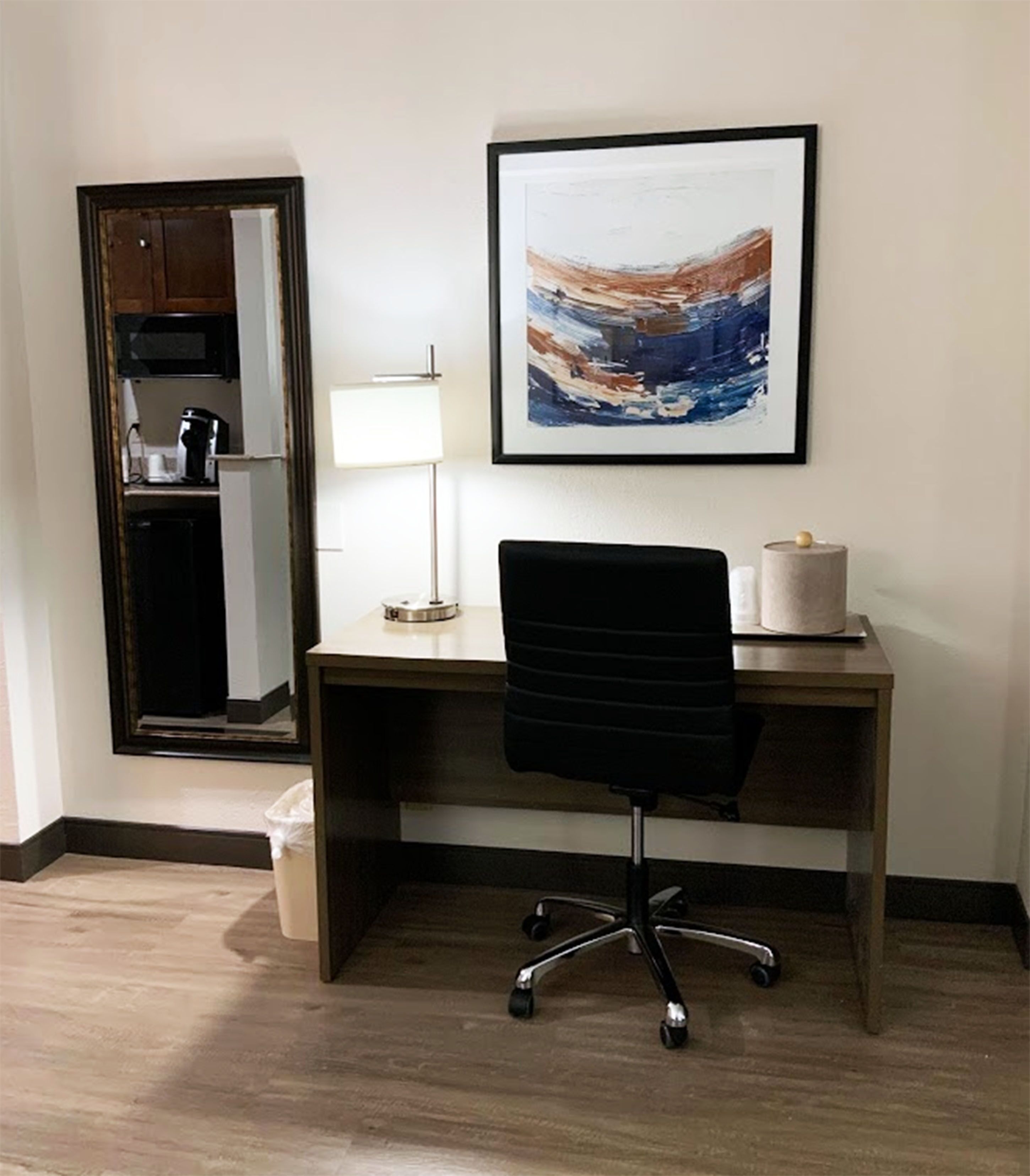 A hotel room with a wooden desk, a black office chair, a framed abstract painting, a lamp, and a microwave.