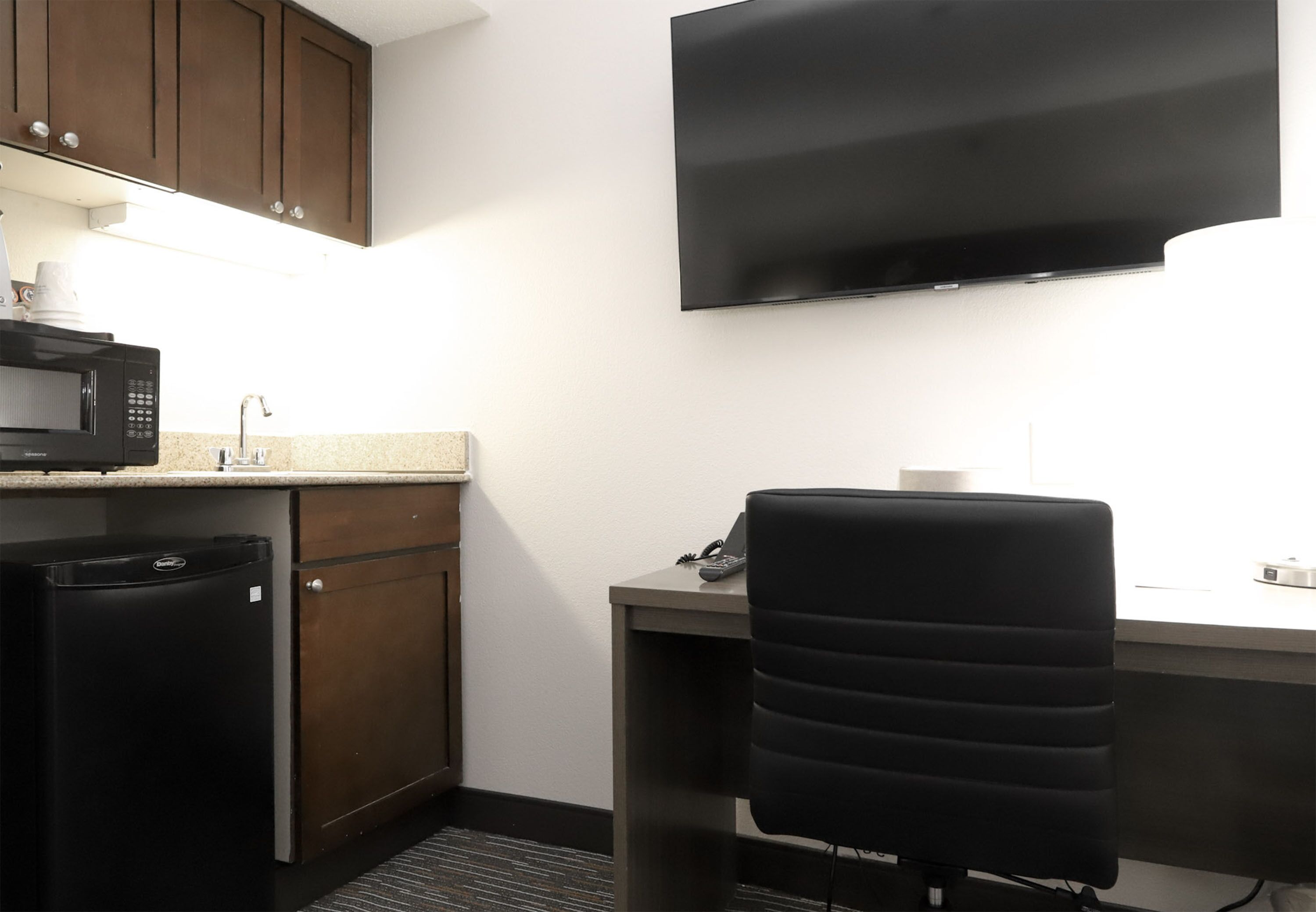 A compact hotel room with a kitchenette, a flat-screen TV mounted on the wall, a desk with a chair, and a small trash bin.