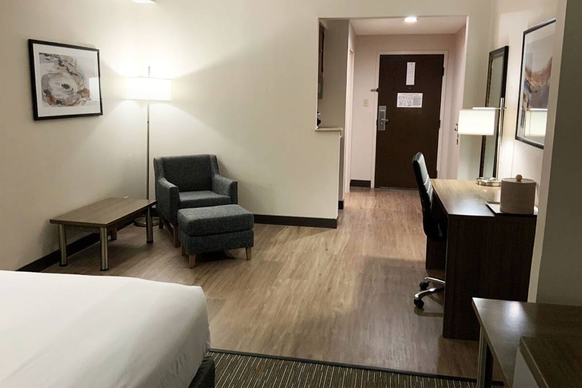A hotel room with a bed, a desk, a chair, a lamp, and a door.