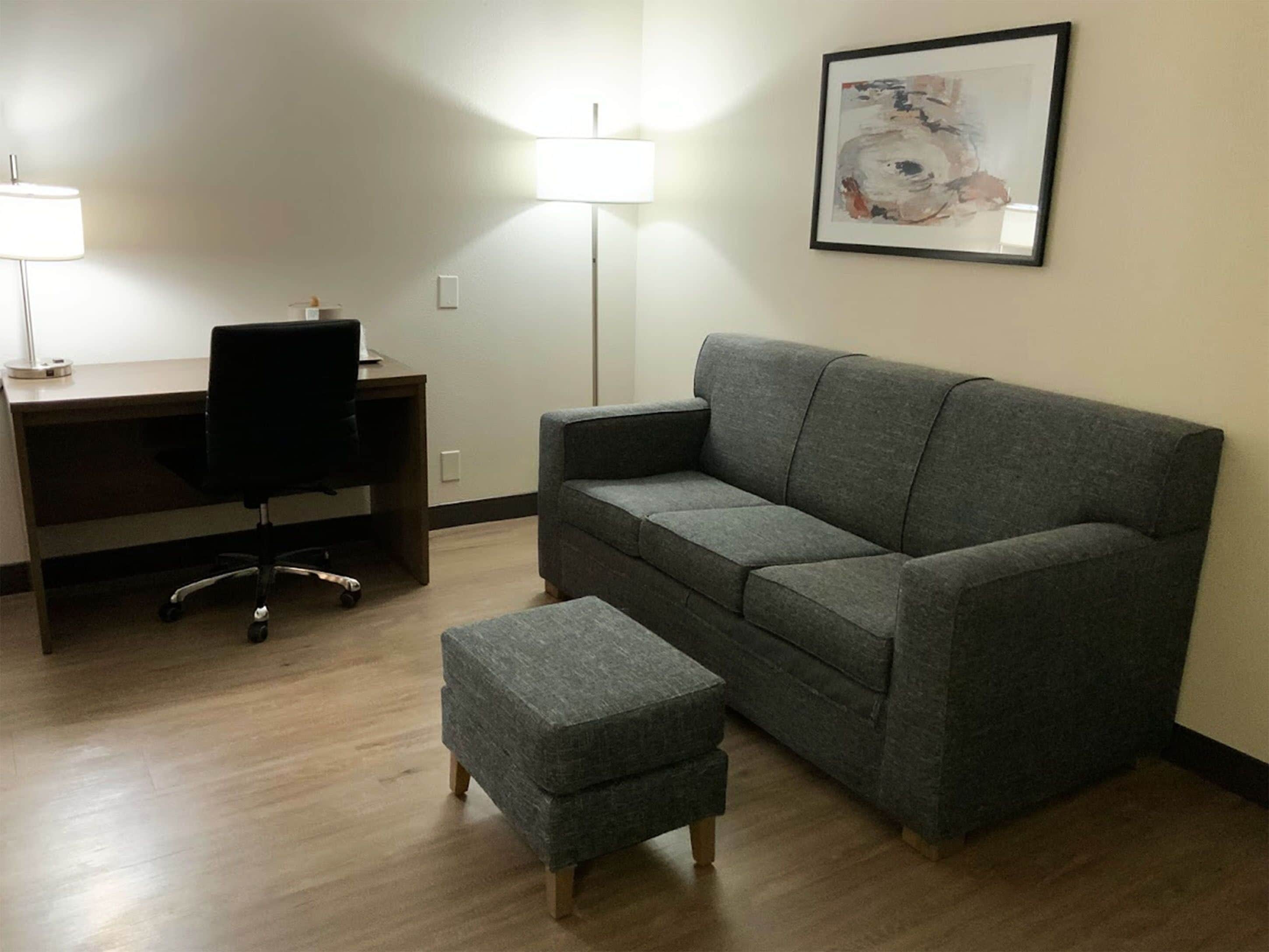 Ein Zimmer mit grauem Sofa, passendem Hocker, Schreibtisch mit Stuhl und Stehlampe.