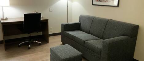 Ein Zimmer mit grauem Sofa, passendem Hocker, Schreibtisch mit Stuhl und Stehlampe.