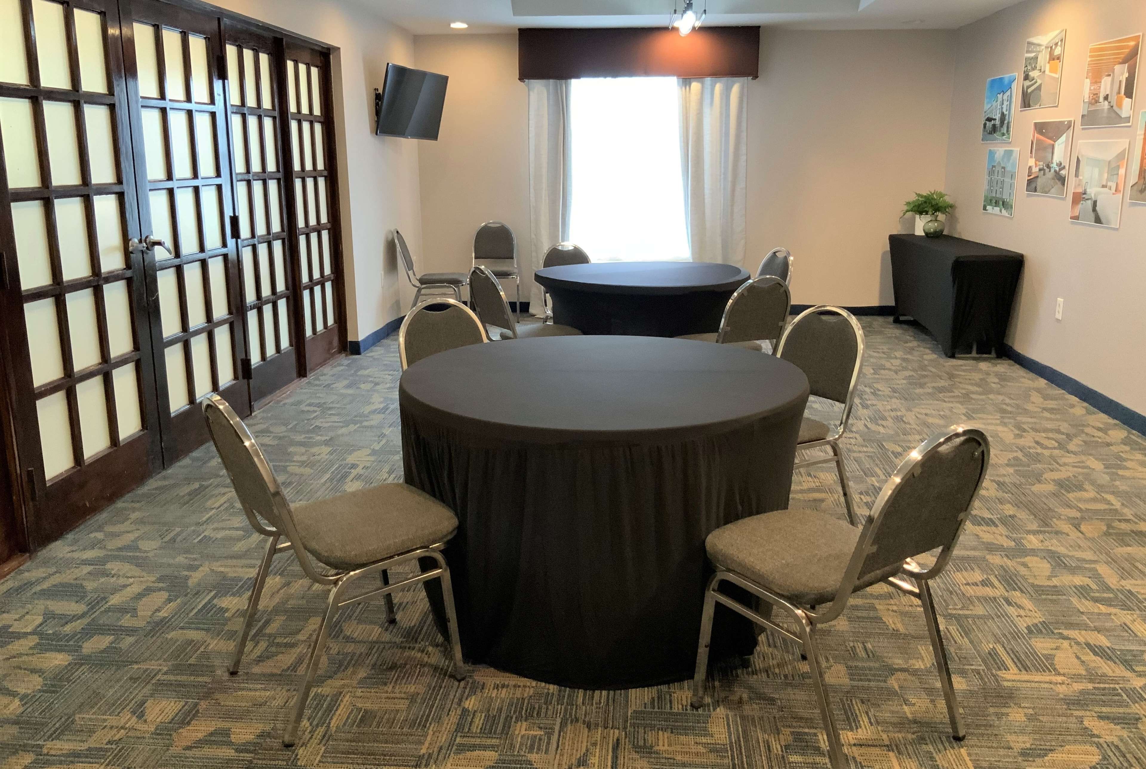 A conference room with a round table, six chairs, a TV on the wall, and a large window with curtains.