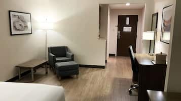A hotel room with a bed, a desk, a chair, a lamp, and a door.