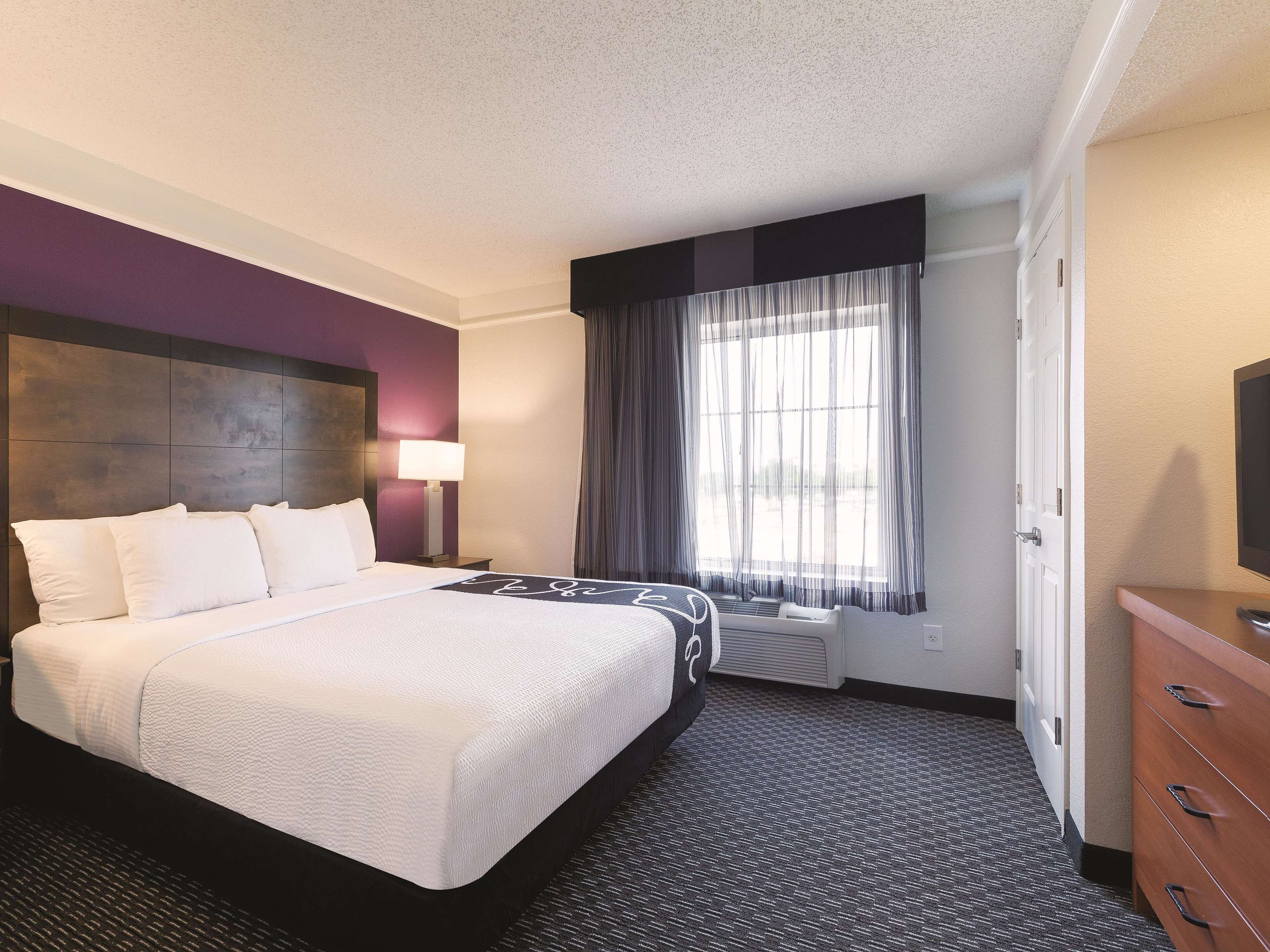 Una habitación de hotel con una cama grande, una mesita de noche con lámpara, una ventana con cortinas y un aparador.