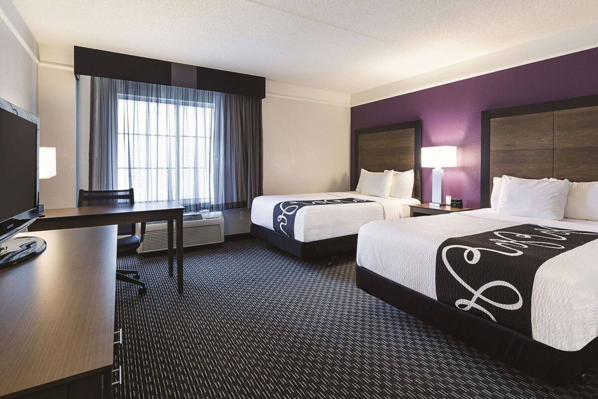 A hotel room with two beds, a desk, a television, and a large window with curtains.