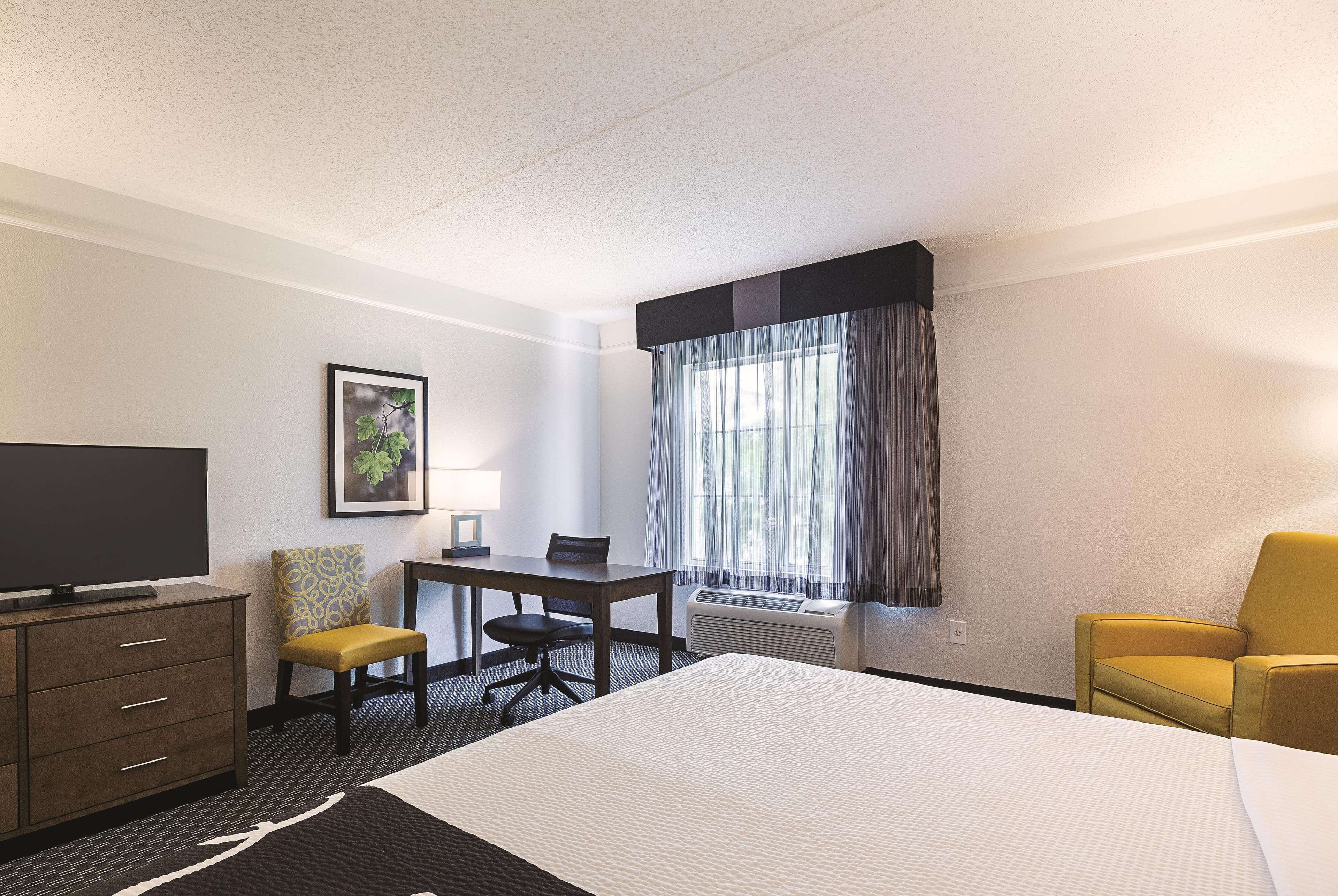 Habitación de hotel con cama, escritorio, silla, televisor y ventana con cortinas.