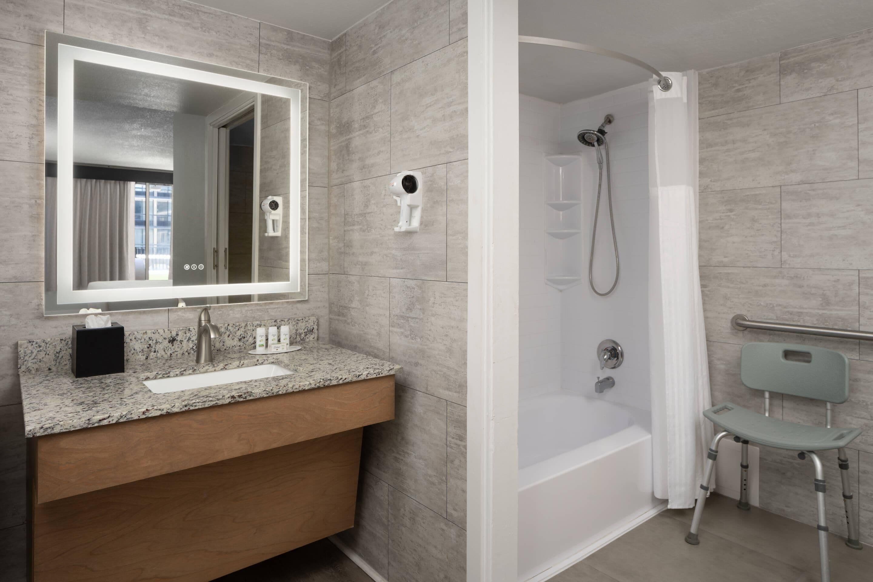 A modern bathroom with a granite countertop, a white sink, a large mirror, a handheld showerhead, a walk-in shower, and a wheelchair-accessible bench.