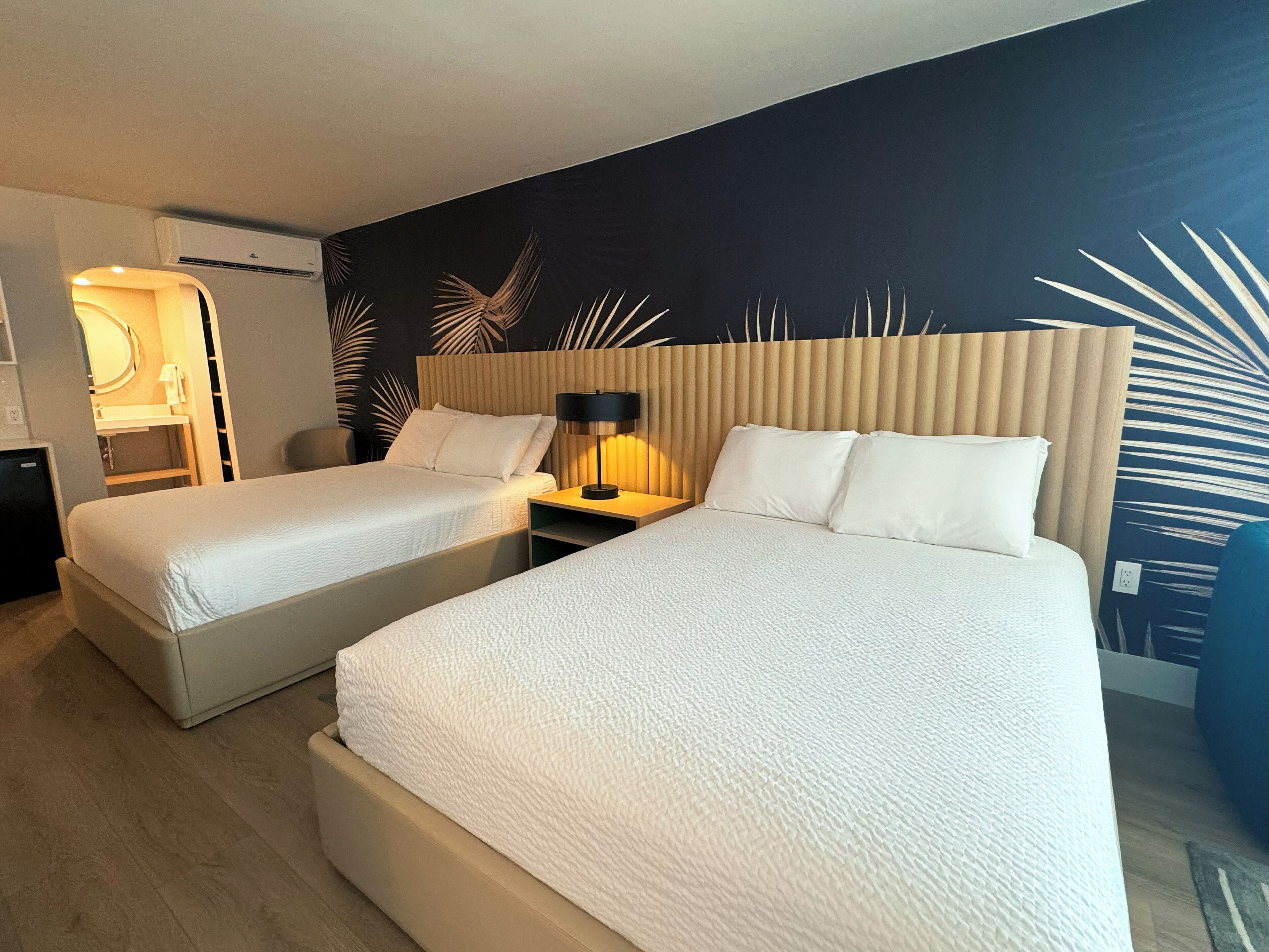 A hotel room with two beds, a nightstand with a lamp, a bathroom with a mirror, and a dark wall with palm leaf patterns.