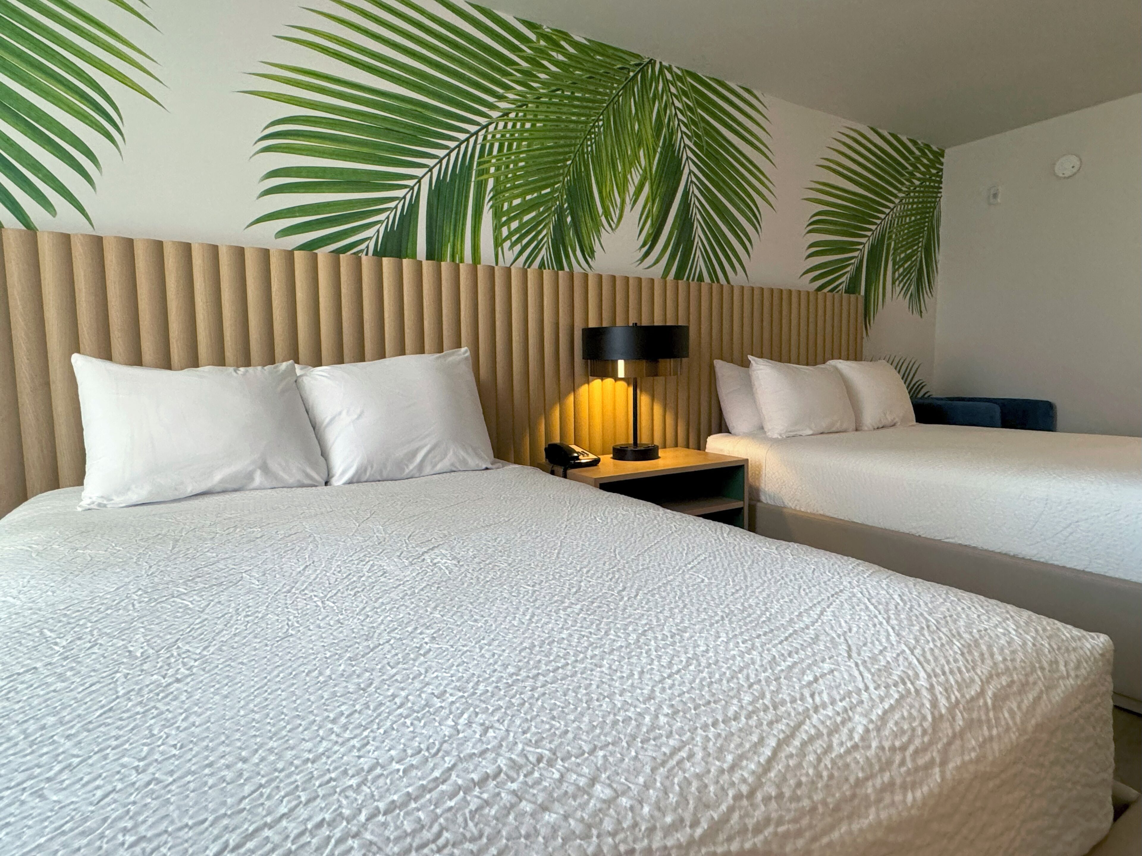 Habitación de hotel con dos camas, un cabecero de madera, una mesita de noche con una lámpara y una pared decorada con motivos de hojas de palma.