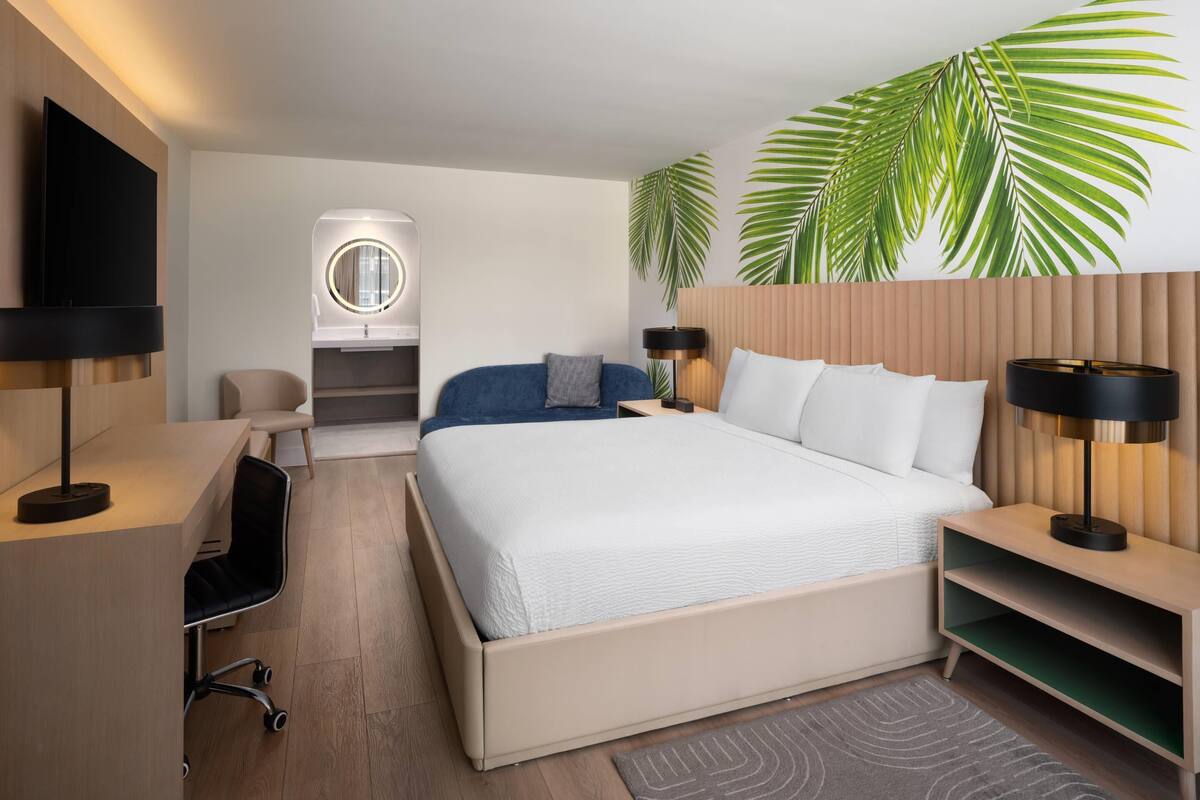 Ein modernes Hotelzimmer mit einem großen Bett, einem Schreibtisch mit Stuhl, einem Fernseher, einem runden Spiegel und einem Wandheizkörper.