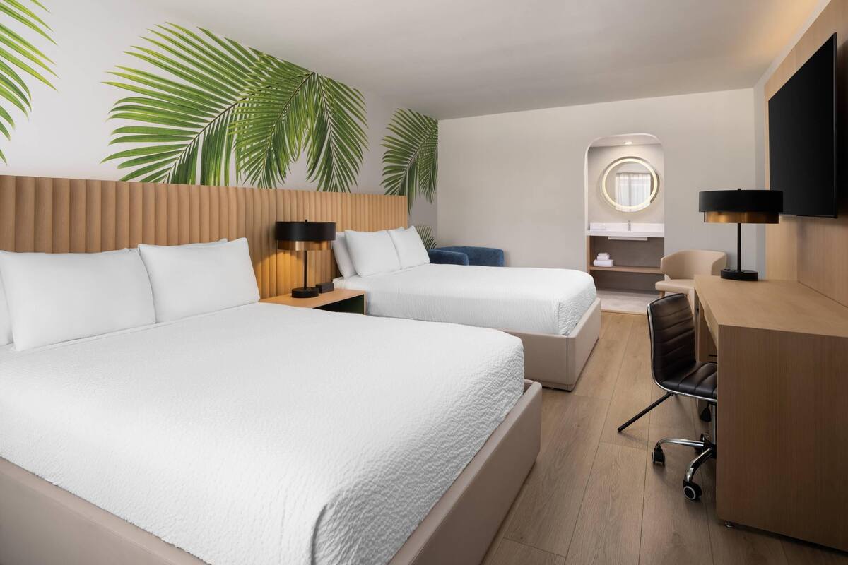 Ein Hotelzimmer mit zwei Betten, einem Schreibtisch, einem Sessel, einem Fernseher und einem Badezimmer mit Spiegel.