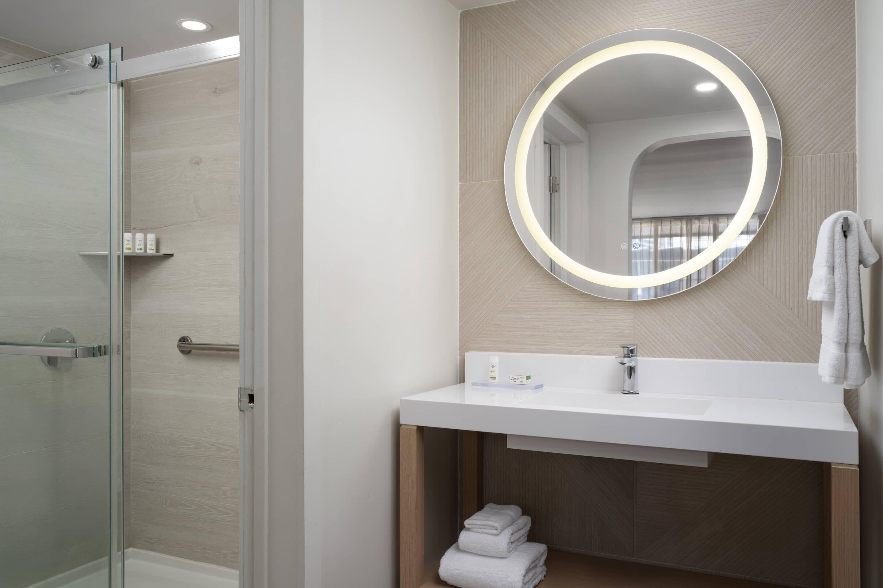 Un baño moderno con cabina de ducha de vidrio, un lavabo blanco con espejo redondo y un perchero para toallas.