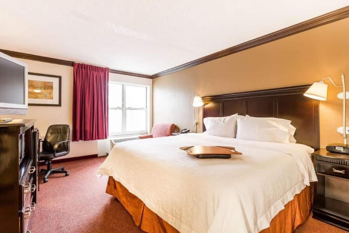 A hotel room with a large bed, a desk, a chair, a lamp, and a window with curtains.