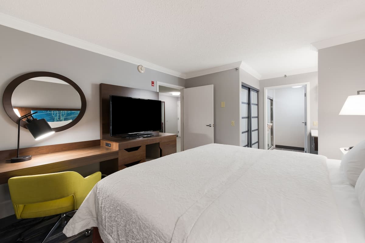 A hotel room with a large bed, a desk with a flat-screen TV, a mirror, and a bright yellow chair.