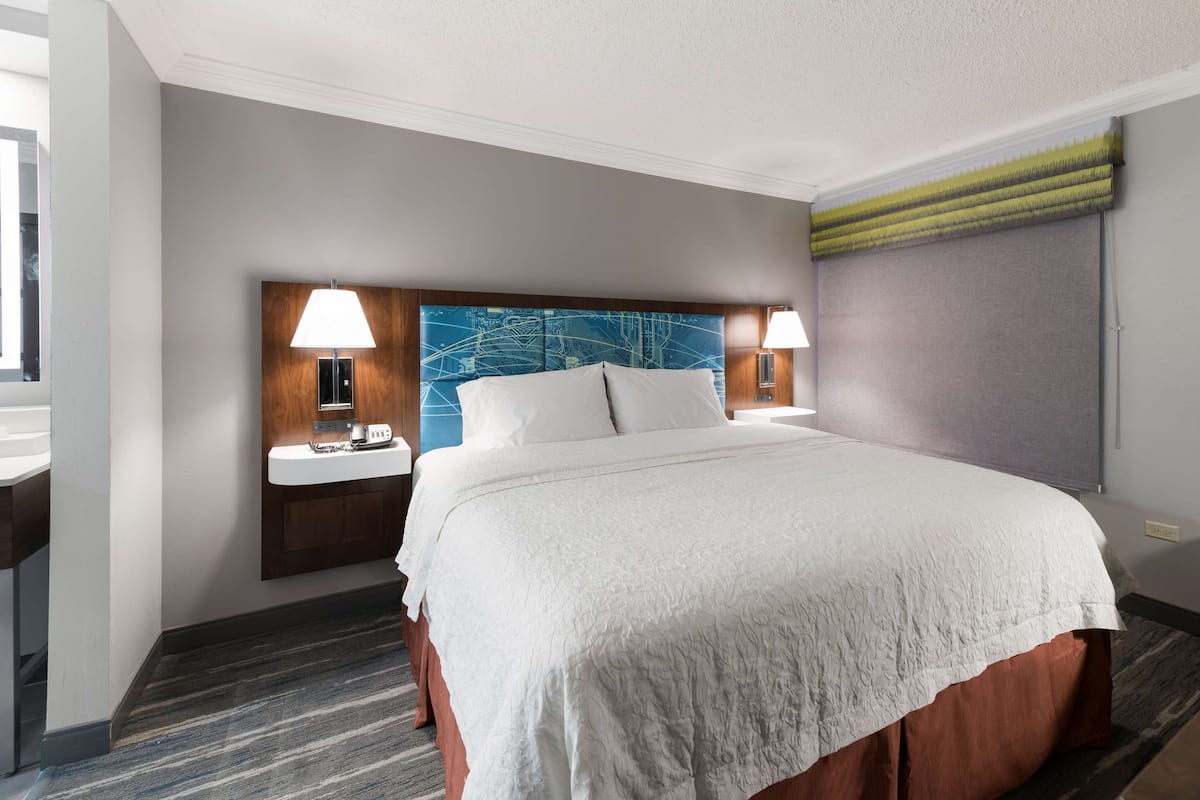 A hotel room with a large bed, two bedside lamps, a nightstand with a lamp, a wall-mounted vanity, and a window with a green blind.