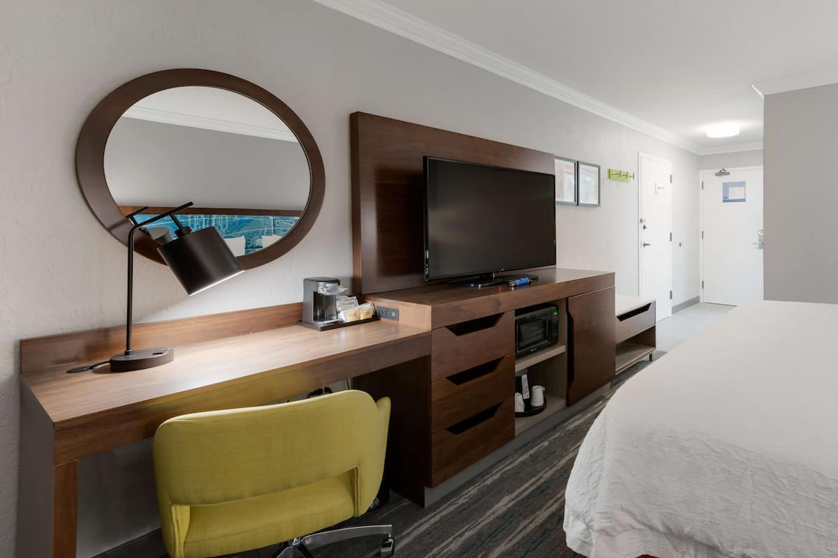 A hotel room with a wooden desk, a television, a yellow chair, a bed, and a round mirror.