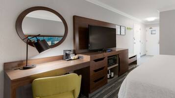 A hotel room with a wooden desk, a television, a yellow chair, a bed, and a round mirror.
