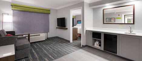 Una habitación de hotel moderna con sofá gris, televisor de pantalla plana, un espejo grande y una kitchenette con lavavajillas.