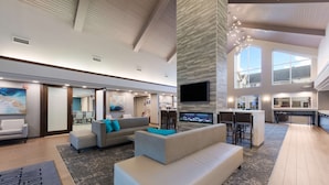 A spacious living area with a large sofa, a coffee table, and a flat-screen TV mounted on a wall. There is a kitchen area in the background with a bar and seating. - Hampton Inn & Suites Chicago/Hoffman Estates (Hoffman Estates)