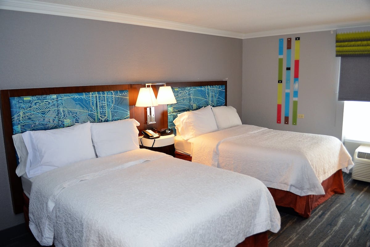 A hotel room with two beds, a nightstand with a phone and lamp, a map on the headboard, and a window with blinds.