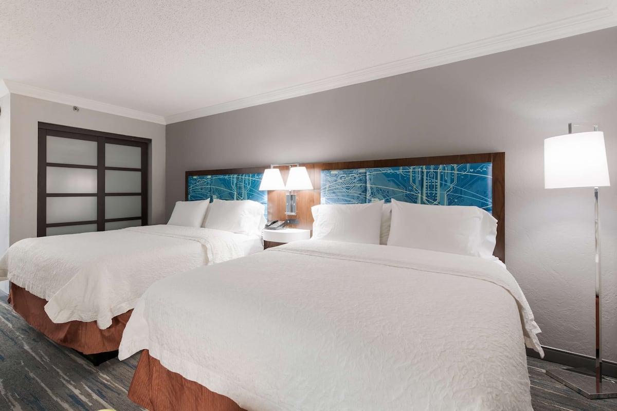 A hotel room with two beds, a large window with a view, a desk with a lamp, and a wall-mounted TV.