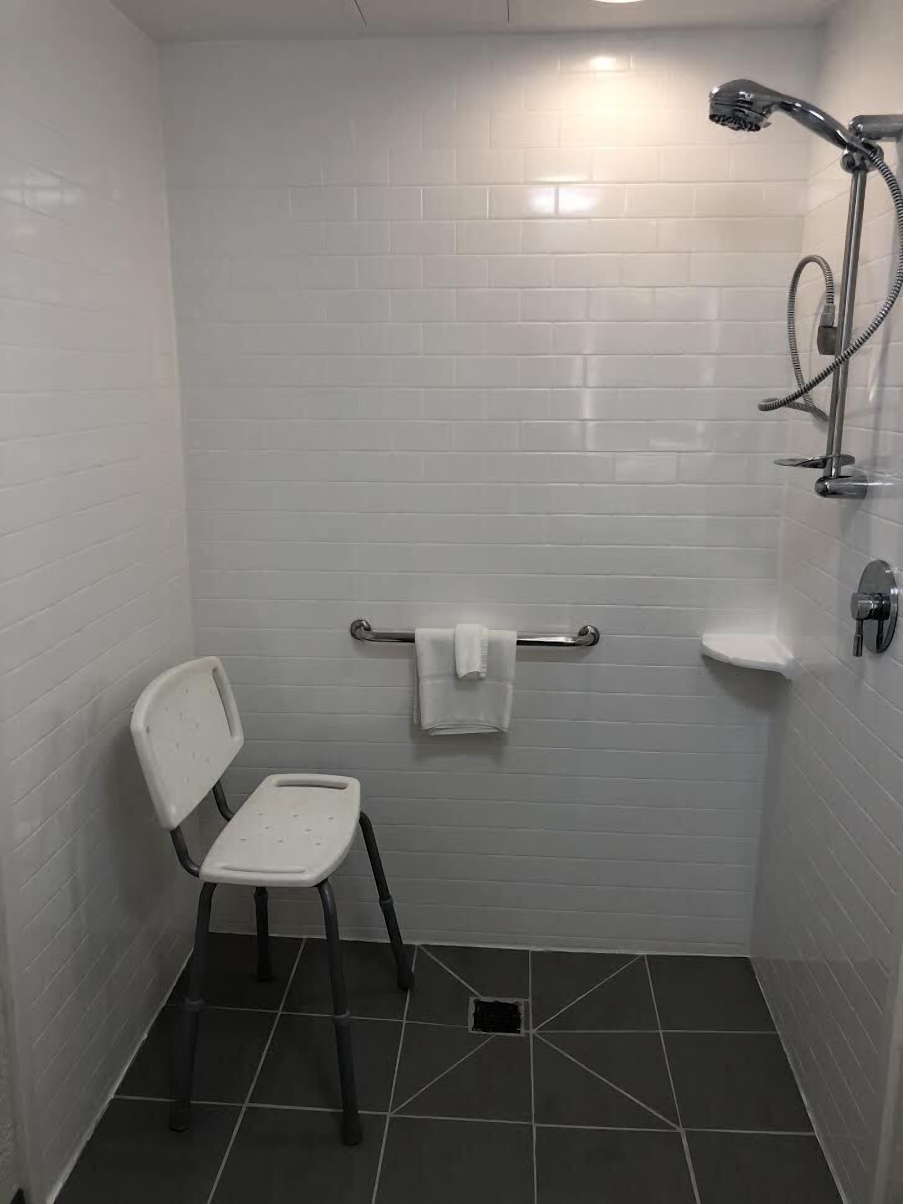 A small, tiled shower area with a white chair, a towel rack, and a handheld showerhead.
