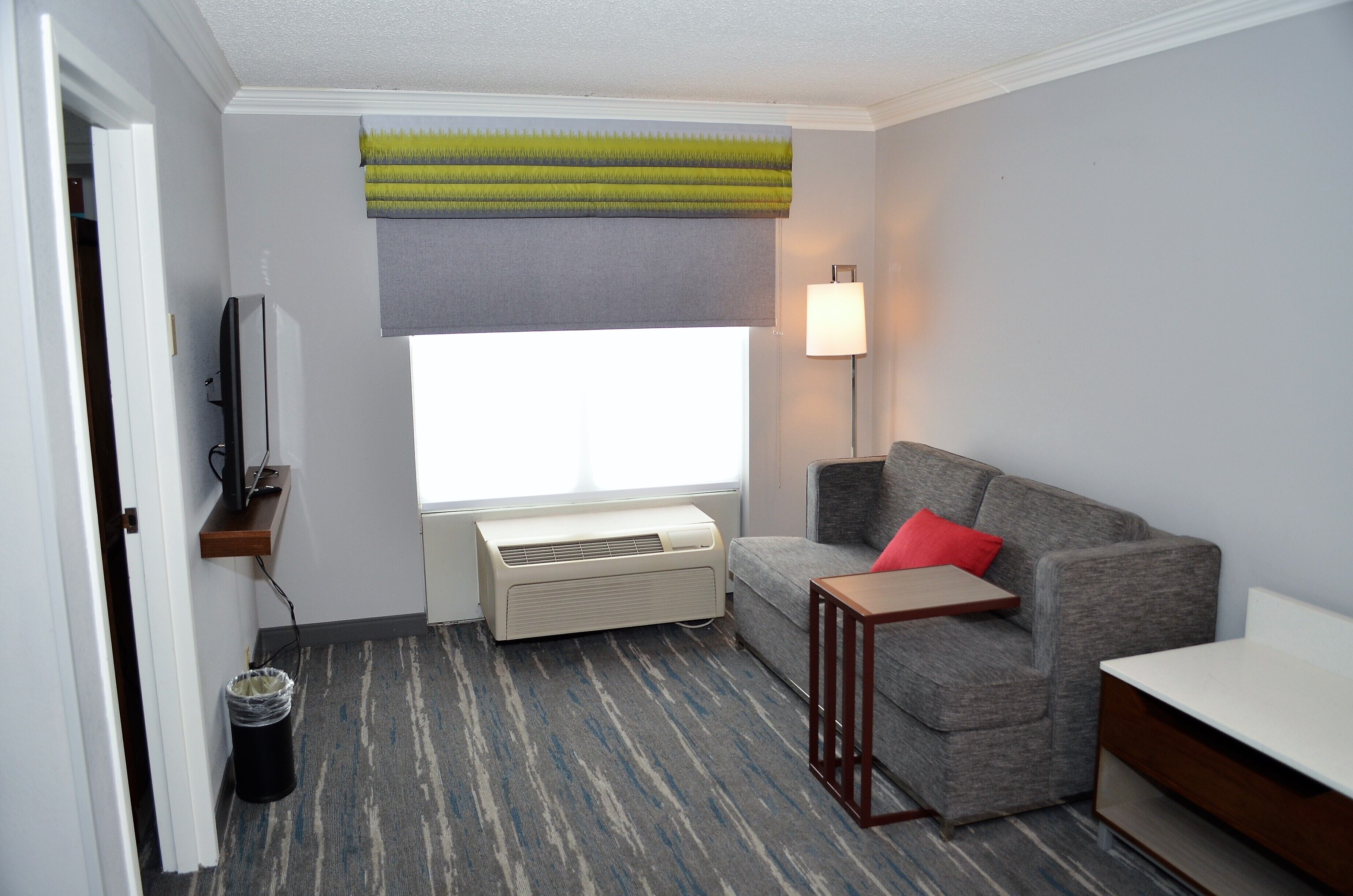 A hotel room with a gray sofa, a small table, a standing lamp, a TV on a shelf, and a window with a green and gray valance.