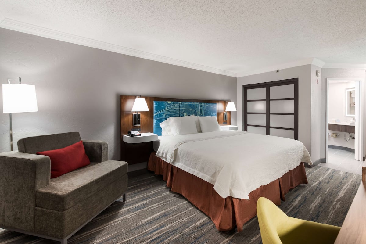 A hotel room with a large bed, a grey sofa with a red pillow, a yellow armchair, a nightstand with lamps, and a bathroom visible through an open door.