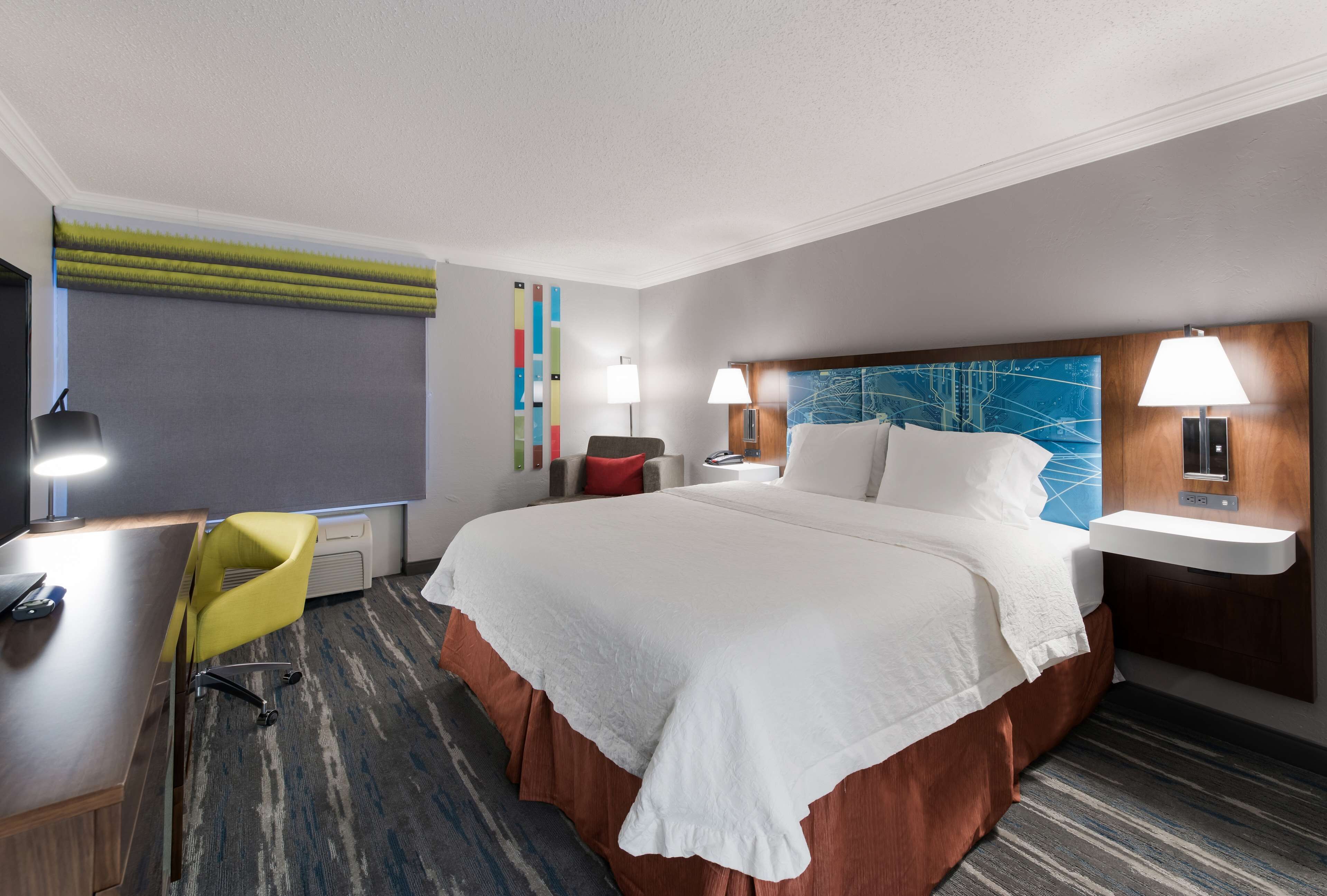 A hotel room with a large bed, a desk with a chair, two bedside lamps, a small table, and a colorful abstract artwork on the wall.