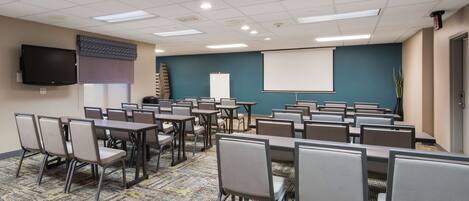 A conference room with multiple tables and chairs, a projector screen, and a television on the wall.