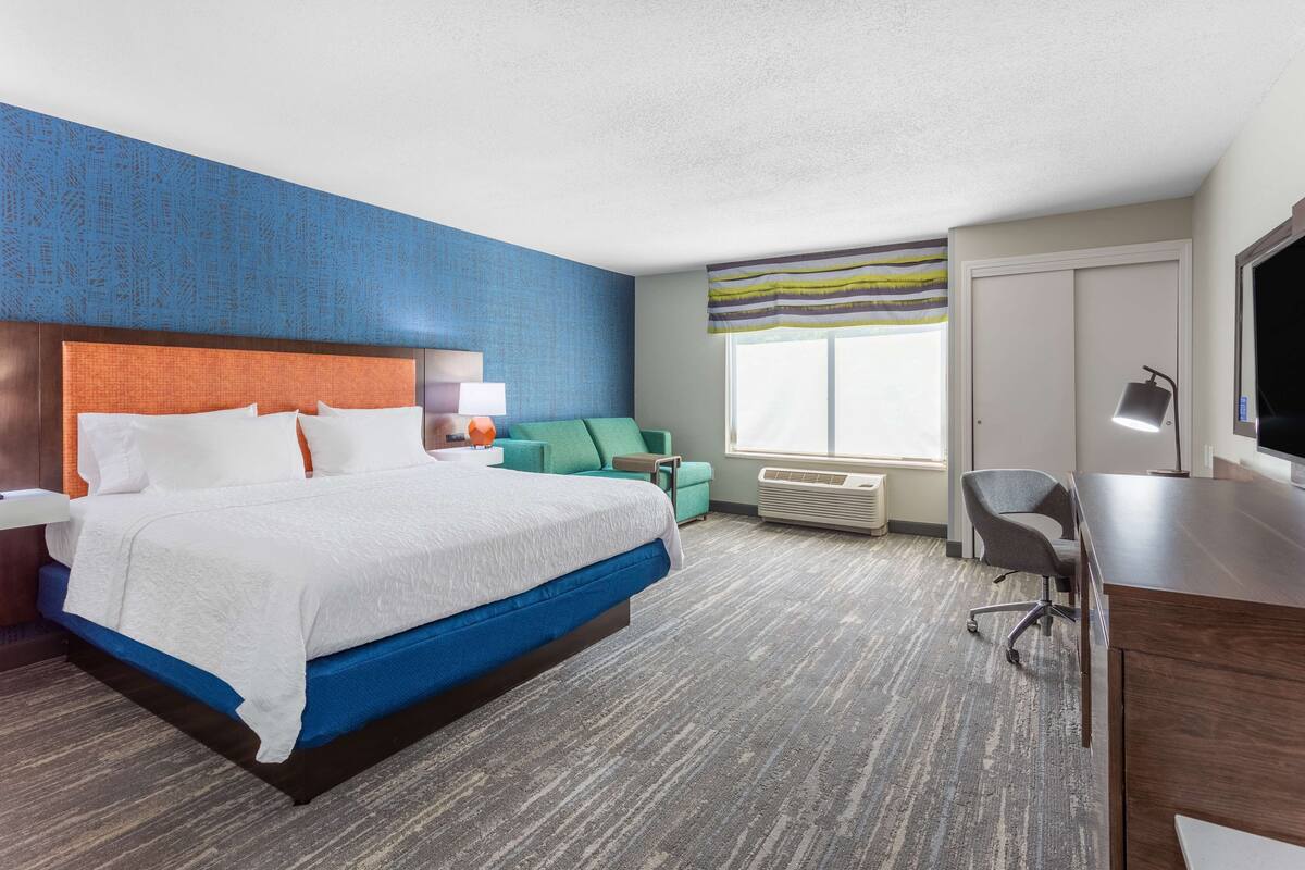 A hotel room with a large bed, a desk, a chair, a TV, and a window with a view.