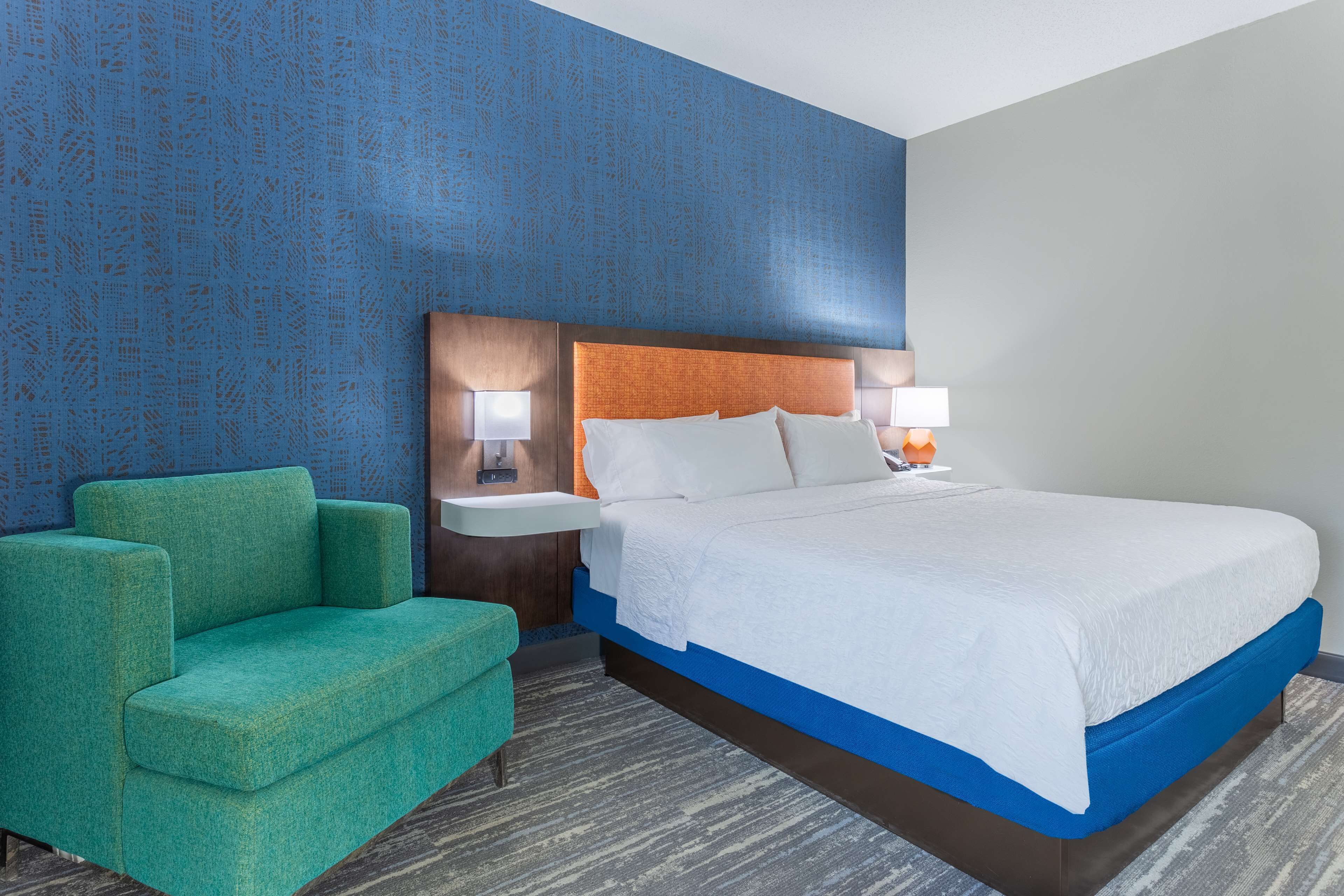 Een hotelkamer met een bed, een groene fauteuil, twee nachtkastjes met lampen en een blauwe muur met een patroon.