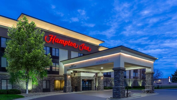 Ein Hampton Inn Hotel mit modernem Design, erkennbar an einer auffälligen roten Signatur, einem überdachten Eingang und Steinsäulen.