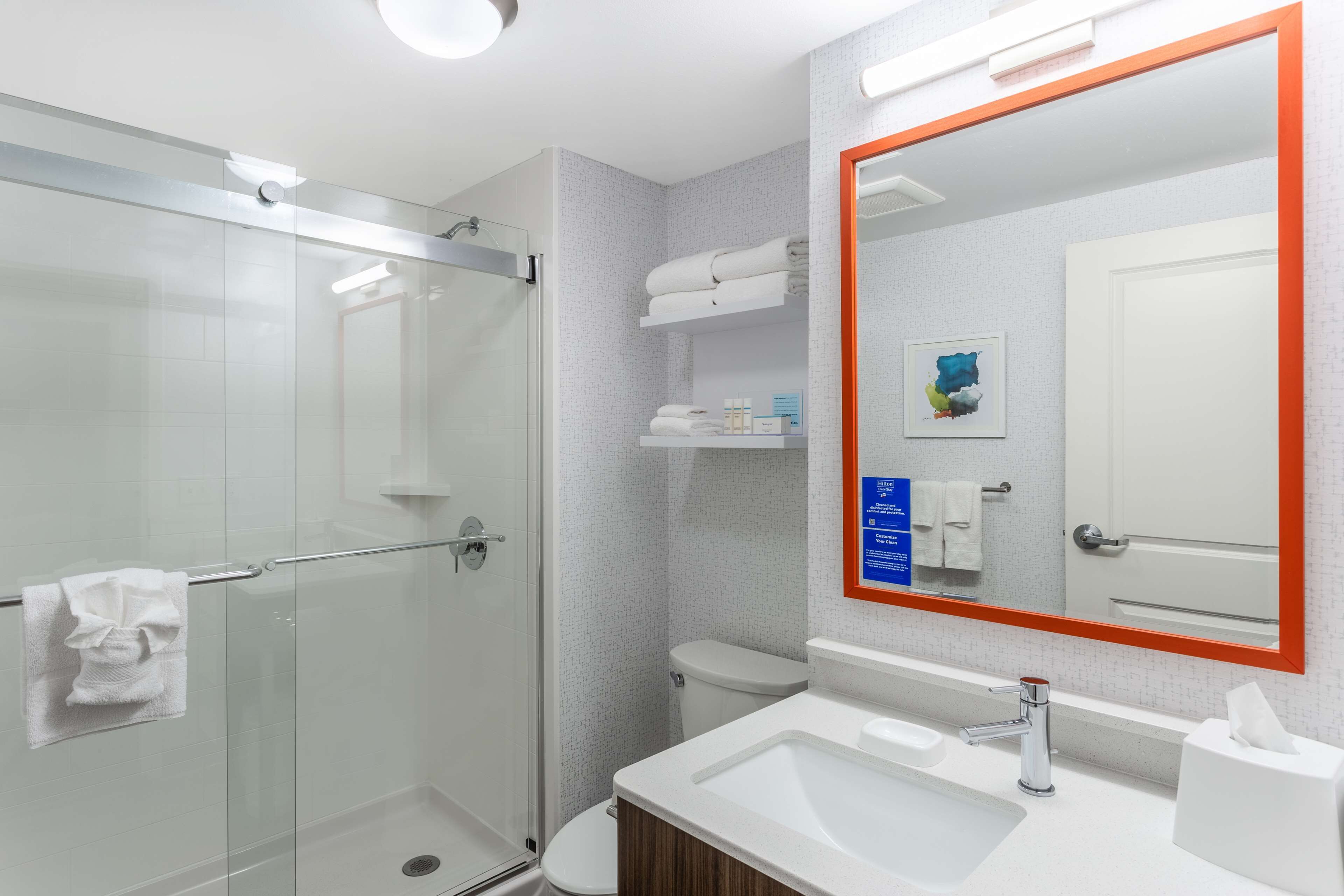 A modern bathroom with a glass shower enclosure, a white sink, and a large mirror with an orange frame.