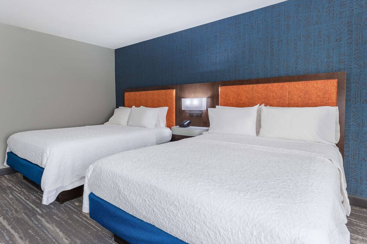 A hotel room with two beds, a nightstand, a lamp, and a wall-mounted TV.