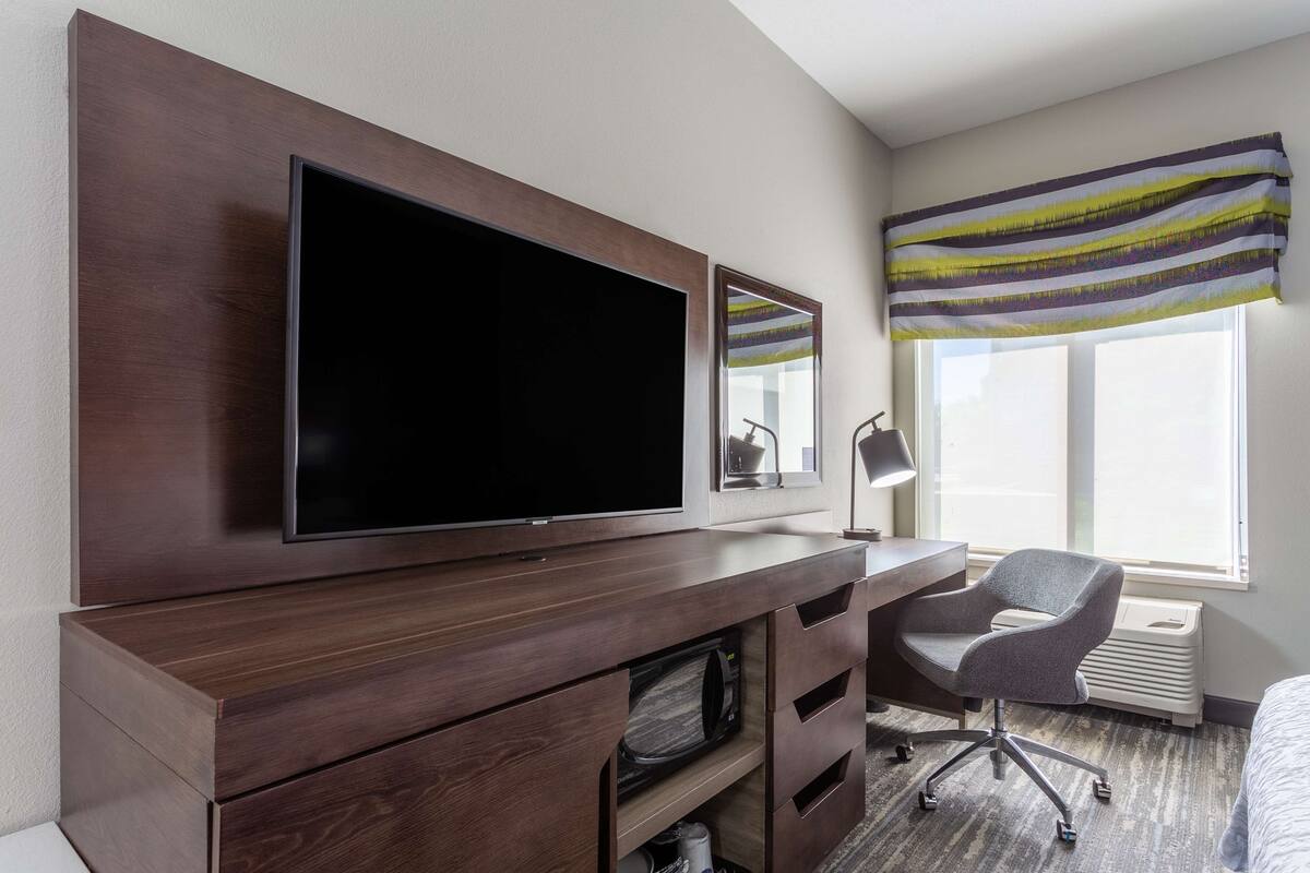 A hotel room with a large flat-screen TV mounted on a wooden cabinet, a desk with a chair, a window with a striped curtain, and a radiator.