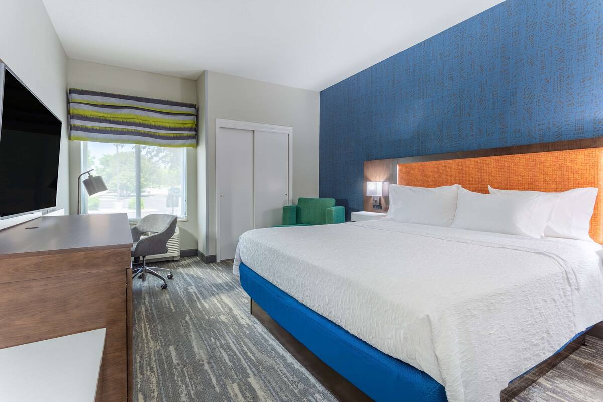 A hotel room with a large bed, a desk with a TV, a chair, and a window with a view of the outdoors.