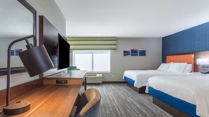 Ein Hotelzimmer mit Schreibtisch, zwei Betten, einem Fernseher und einem Fenster mit gestreiftem Rollo.