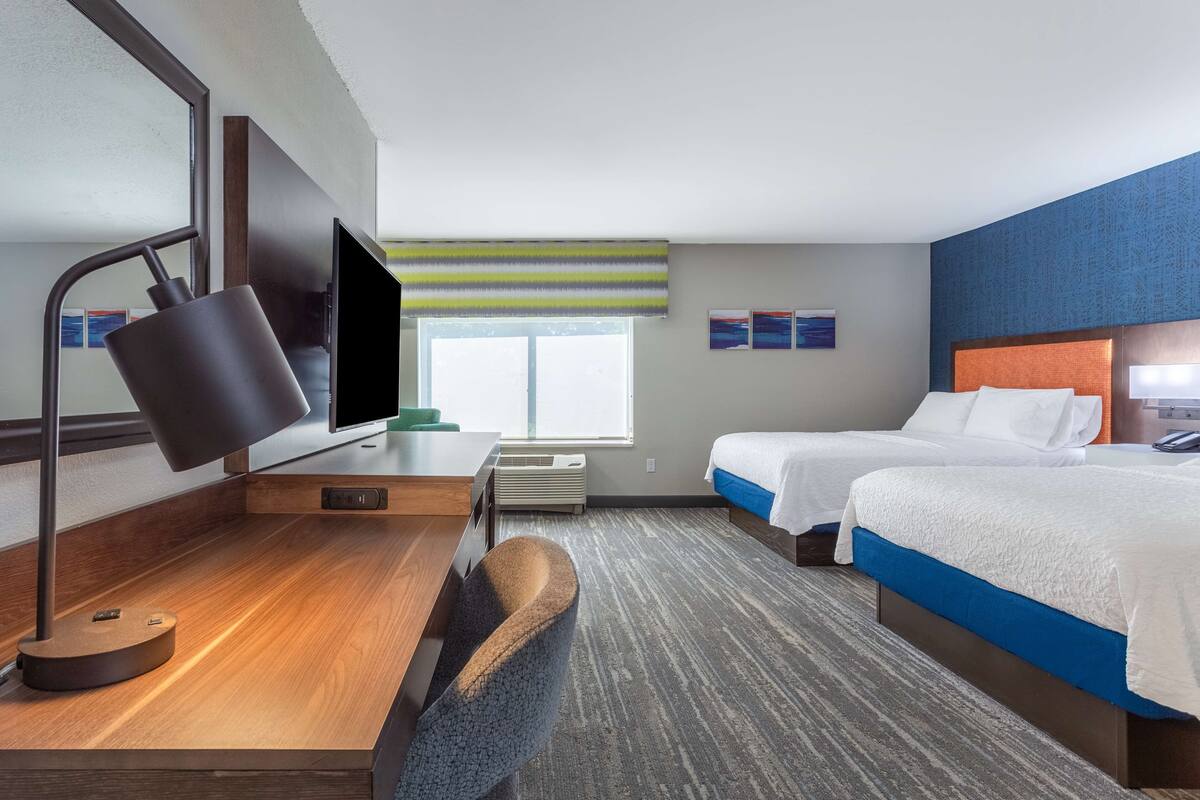 A hotel room with a desk, two beds, a TV, and a window with a striped blind.