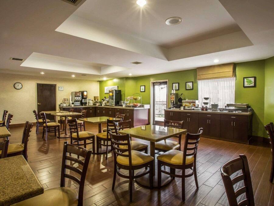 A hotel dining area with multiple tables and chairs, a kitchen area with a green wall, and a clock on the wall.