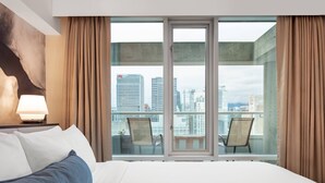 Ein Hotelzimmer mit einem großen Bett, einem Nachttisch mit Lampe, Blick auf die Stadt durch ein großes Fenster und einem Balkon mit Sitzgelegenheiten.